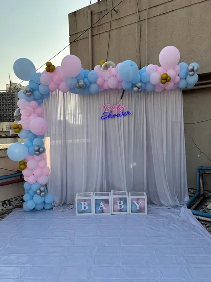 A baby shower photo backdrop set up on a terrace. This features a pastel pink and blue balloon arch, a sheer white curtain, and transparent 'BABY' boxes.