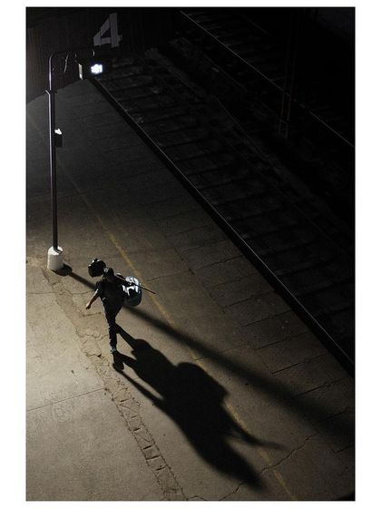 An overhead shot of a lone traveler walking on a train platform at night. The single light source casts a long, dramatic shadow, creating a feeling of solitude and journey.