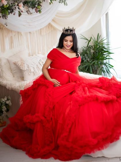 A queen on her throne. This mom-to-be is seated on our studio bed setup, draped in a magnificent red gown and wearing a crown, creating a portrait of pure elegance.