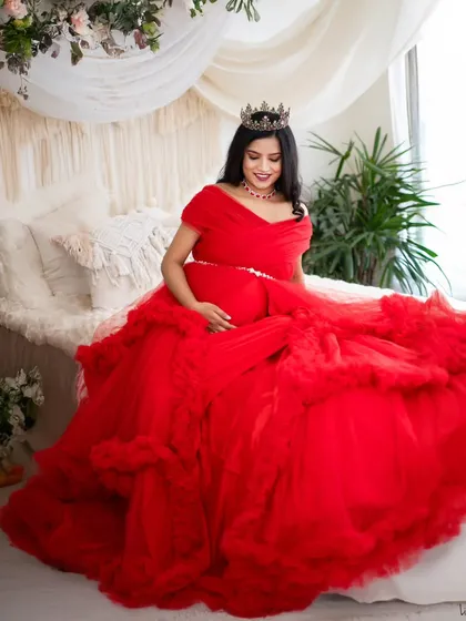 A queen on her throne. This mom-to-be is seated on our studio bed setup, draped in a magnificent red gown and wearing a crown, creating a portrait of pure elegance.