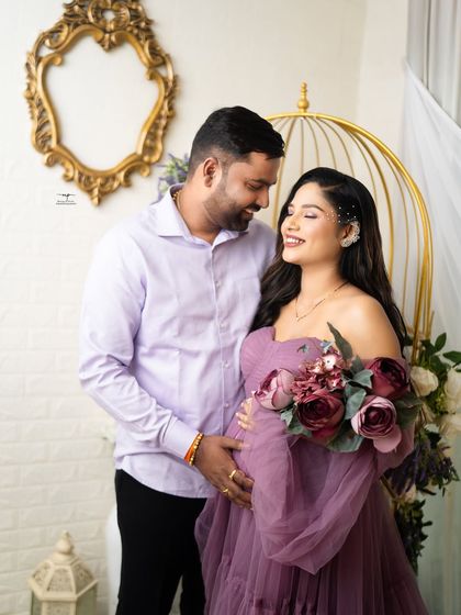 A smiling couple shares a look of love during their purple-themed maternity shoot. The soft lighting and floral arrangements complement the dreamy, romantic mood.