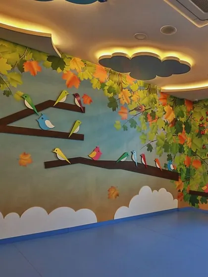 This classroom is inspired by a forest canopy, with colorful birds perched on tree branches painted on the walls. The ceiling features cloud-shaped light fixtures, bringing the feeling of being outdoors inside.