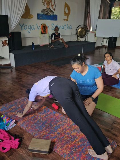 A teacher assists a student in Downward-Facing Dog, ensuring her shoulders are properly engaged and her spine is long.
