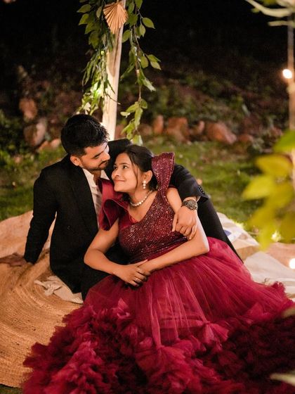 A cozy and romantic pre-wedding photo. The couple is seated in a beautifully styled night setup, with the bride's red ruffled gown creating a stunning visual.