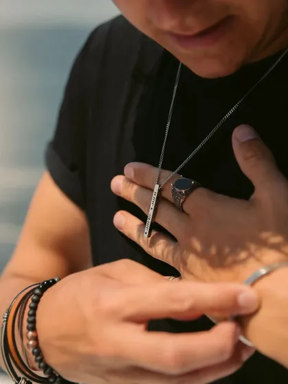 A man adjusting his silver-toned bar pendant, shown here with other accessories like rings and bracelets. This highlights the pendant as part of a complete, styled look.