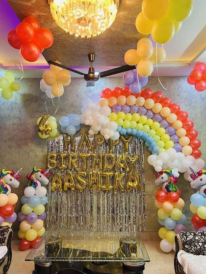 A rainbow and unicorn theme decoration for a living room, featuring a large rainbow balloon arch against a silver fringe curtain.