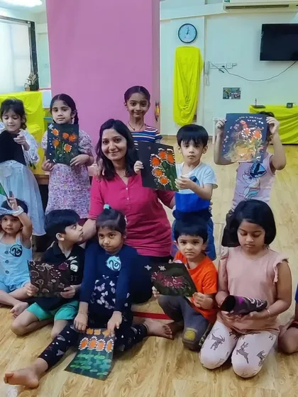 A happy group photo from a summer art camp. I'm sitting on the floor with a large group of young children, all holding up the floral paintings they created on black paper.