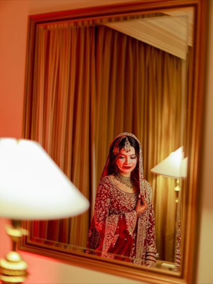 An artistic mirror reflection of the bride. The warm, reddish lighting creates a romantic and dramatic mood, highlighting her classic red bridal look.