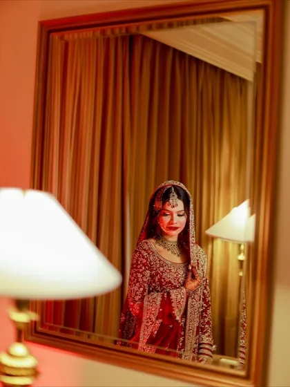 An artistic mirror reflection of the bride. The warm, reddish lighting creates a romantic and dramatic mood, highlighting her classic red bridal look.