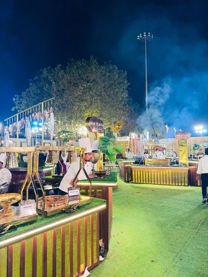A wide-angle view of a bustling outdoor corporate event at night. Multiple food stations, including our rustic wooden and modern gold counters, showcase the scale and variety I can bring to any large gathering.