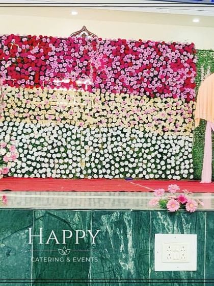 A large-scale stage design combining a full green wall with a textured floral panel in shades of red, pink, and white. The spiral floral accent on the side adds a unique, artistic touch to this reception backdrop.