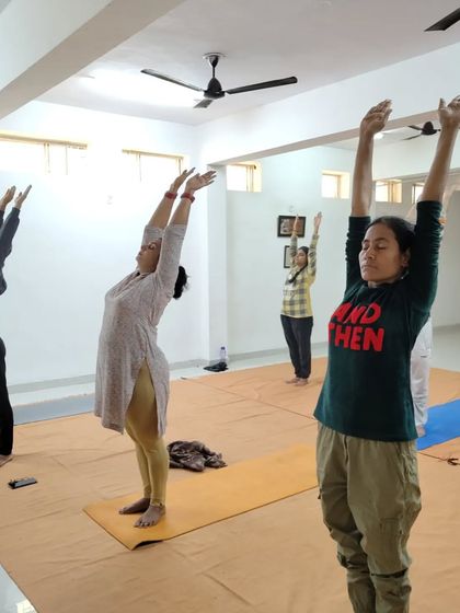 A group of women reach upwards in a backbend during a Hindi Surya Kriya session.