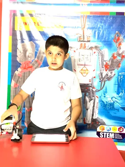 A young boy at a red table with his LEGO robot and tablet, ready for a programming session.