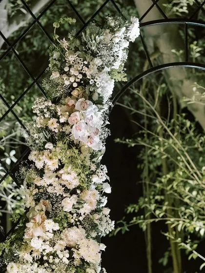 A close-up of a floral arch, showing the intricate weaving of white orchids, roses, and baby's breath on a dark trellis frame.