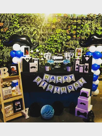 The full 'Little Man' themed dessert table and decor for a third birthday party. This setup includes mustache themed balloon columns, custom banners, and a personalized prop shelf.