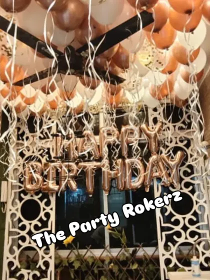 A surprise birthday setup in a room with rose gold and white balloons on the ceiling and "Happy Birthday" foil letters on the wall.