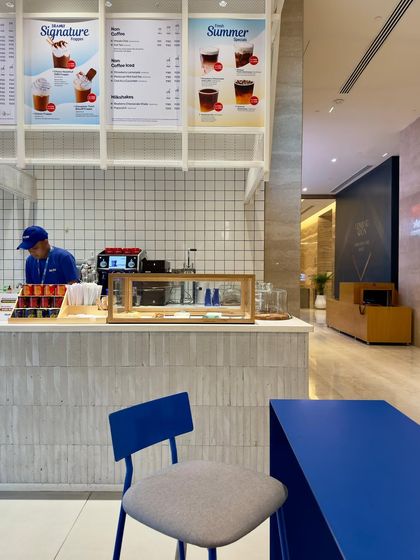 The view from the seating area towards the service counter at Beanly. The design uses clean white tiles and a simple counter to create an efficient and organized QSR feel, while the blue chair in the foreground ties the space together.