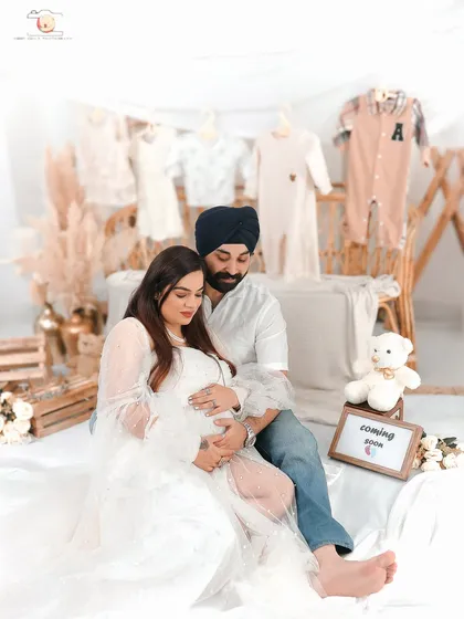 A tender moment between expectant parents during a studio maternity session. The soft, neutral backdrop with hanging baby clothes and a 'Coming Soon' sign creates a feeling of gentle anticipation and love.