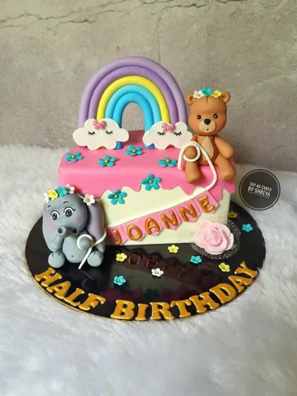 Celebrating a half birthday is twice the fun. This adorable cake marks the six-month milestone with cute, handmade elephant and teddy bear figurines holding a "Half Birthday" banner under a sweet pastel rainbow.