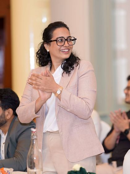 A candid shot of me applauding speakers at a corporate event. Dressed in a smart pink blazer, I am actively listening and engaged with the program's content.
