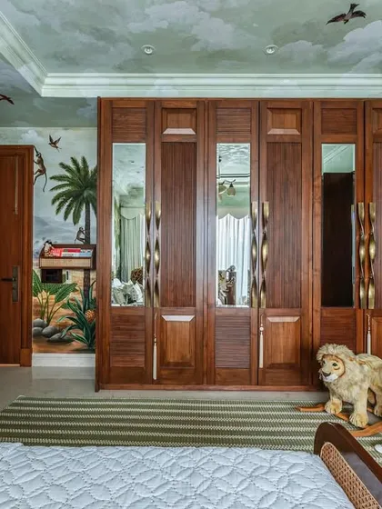 This view showcases the custom wooden wardrobes with mirrored panels, which reflect the beautiful jungle mural on the opposite wall. Every element, including the stuffed lion, adds to the room's playful explorer theme.