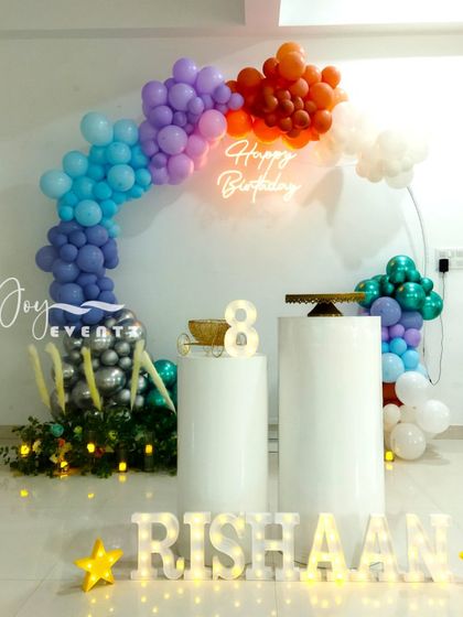 This beautiful balloon ring decoration for an 8th birthday uses a rainbow of colors, from deep blues and purples to orange and white. The setup is completed with a neon sign, cake plinths, and illuminated marquee letters for the name 'RISHAAN'.