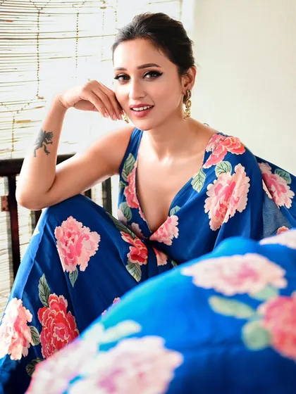 A full-body shot showing the beautiful floral print of the blue saree. The relaxed, seated pose is both graceful and modern.