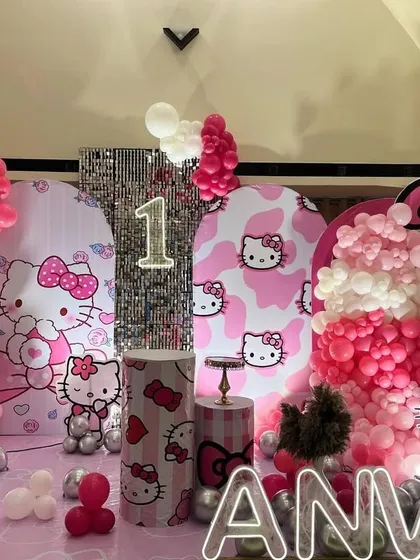 A cute Hello Kitty themed first birthday party. The setup includes multiple backdrops with Hello Kitty prints, pink and white balloon columns, and a personalized light-up name sign.