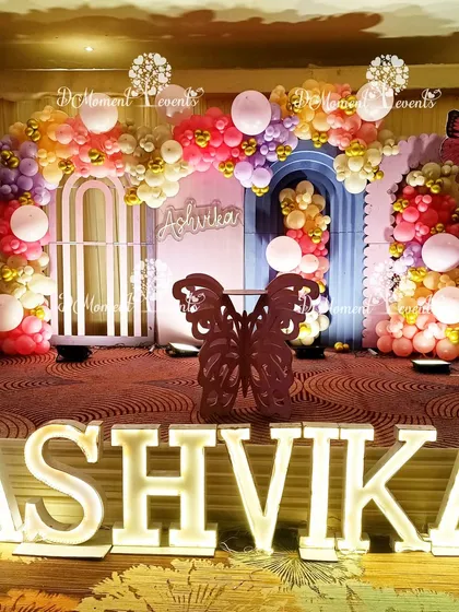 A vibrant stage for Ashvika's welcome party, featuring a fairy and butterfly theme. The decor includes colorful balloon garlands, marquee letters, and butterfly props against pink and blue arched backdrops.