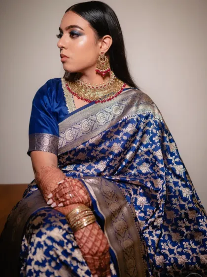 Seated portrait of Sonia showcasing her complete reception look. The rich blue of her saree and the dramatic eye makeup are balanced by a flawless base and nude lips, creating a look of pure sophistication.