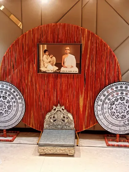 A unique backdrop for a Jain function featuring a large circular element made of red threads. The setup includes a traditional silver chair (singhasan) and Pichwai-style painted discs.