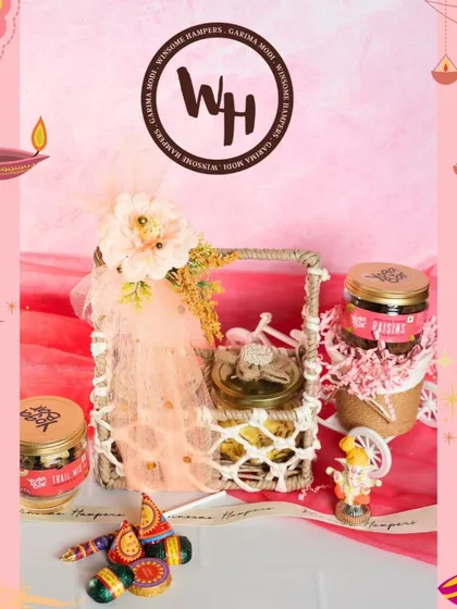 A charming and traditional Diwali gift basket. This hamper includes jars of trail mix and raisins, a small Ganesha figurine, and chocolate crackers, all presented in a rustic woven basket with delicate net and floral wrapping.