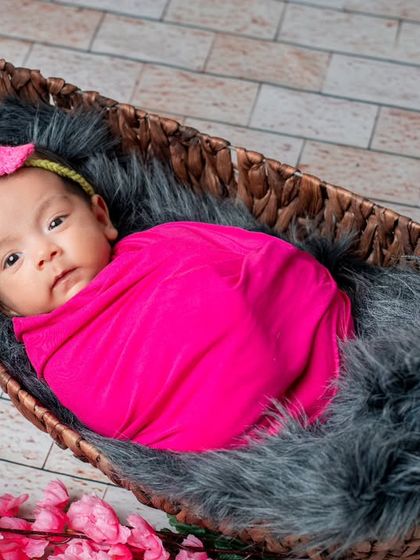 This baby girl is wrapped in a bright pink swaddle, nestled comfortably in a basket. The pop of color against the neutral fur and flooring creates a vibrant and eye-catching portrait.