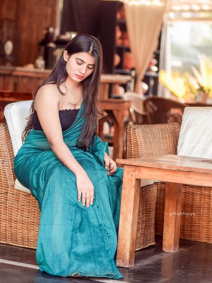 A modern take on saree styling. The model wears a teal saree paired with a contemporary black top, shot in a cafe with earthy tones. This shows how traditional wear can be styled for a modern, chic look.