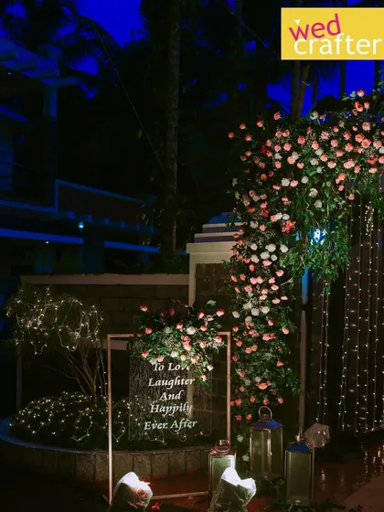 A romantic corner at night, featuring a lit sign that reads "To Love, Laughter And Happily Ever After" surrounded by flowers and fairy lights.