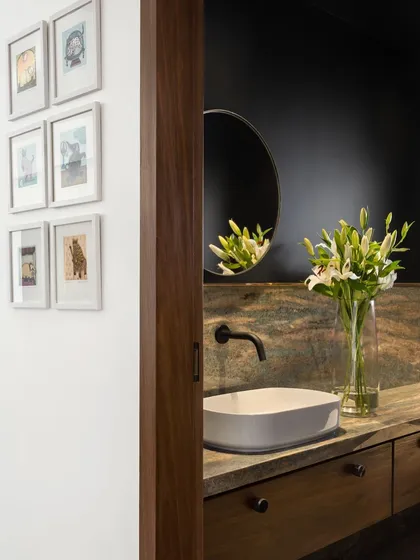 In the West Coast Residence, a powder room is revealed behind a sliding wooden door. The dark, moody walls create a dramatic backdrop for the veined stone countertop and simple vessel sink.