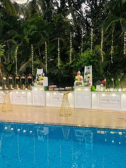 A beautiful poolside catering setup for a 75th anniversary celebration. The clean white food counters are decorated with fairy lights that reflect in the pool, creating a serene and magical ambiance for the evening.