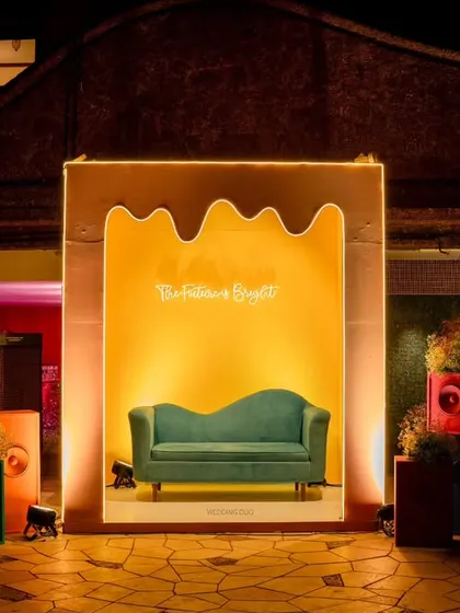 "The Future is Bright" photo booth. A comfortable teal sofa is set against a bright yellow, organically shaped backdrop, flanked by colorful speaker installations.
