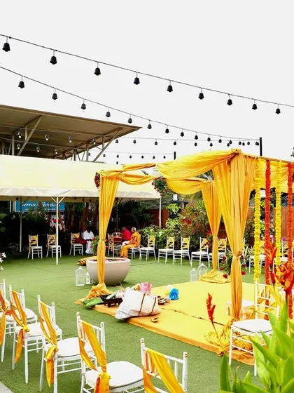 A wide view of a Haldi ceremony setup, with string lights overhead and lush greenery surrounding the space.