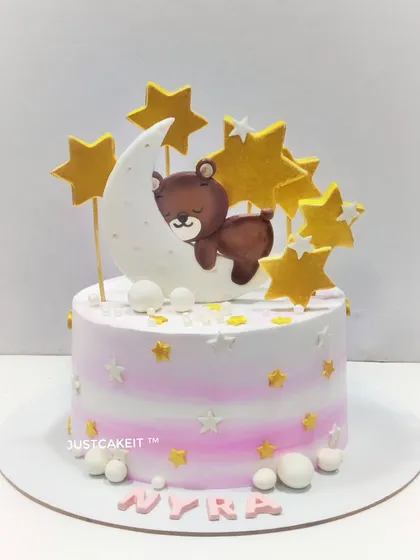 A dreamy cake设计 featuring a teddy bear sleeping on the moon. The soft pink and white ombre and golden stars make it perfect for a baby's celebration.