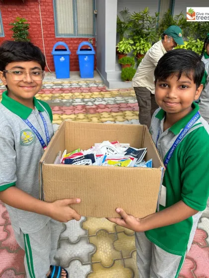 Two students hold a large box filled with collected milk packets. This simple act of recycling provides us with thousands of free, durable nursery bags.