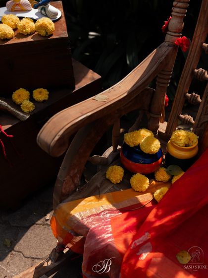 A close-up of the antique chair installation, showing marigold flowers scattered on a vibrant red and orange dupatta. The hand-painted pickle jars add another layer of quirky, upcycled charm.