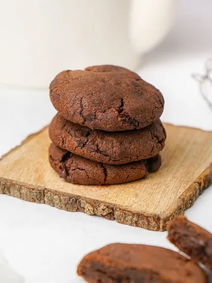 A stack of soft, dark chocolate cookies on a small wooden board. The clean, bright background makes the rich color of the cookies stand out.