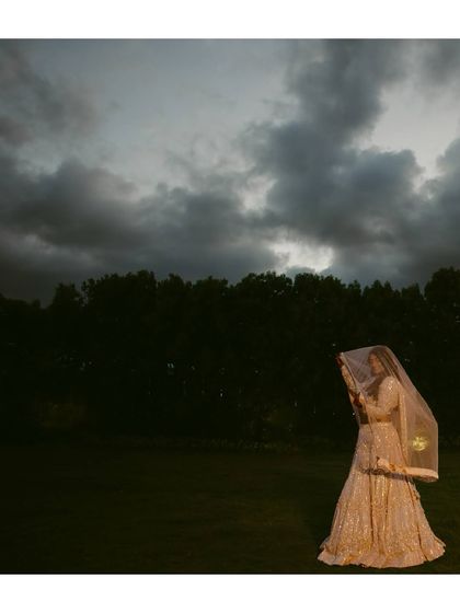 A bride in a sparkling lehenga stands in a field under a dramatic, cloudy sky. This wide shot creates a sense of fairytale magic.