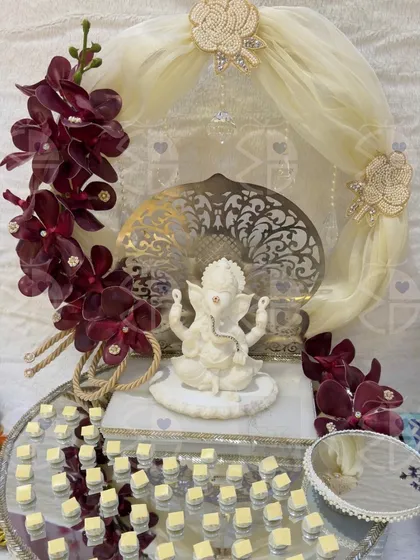 A 'Shaadi Ka Shagun' hamper designed to invoke blessings. A serene white Ganesha idol sits at the center, surrounded by a platter of sweets and framed by deep red orchids, creating a divine and luxurious offering.
