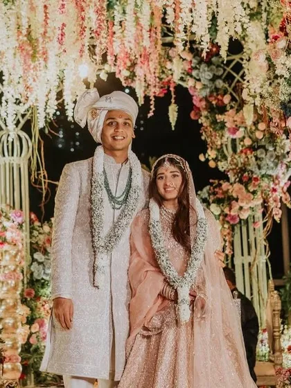 A happy couple poses after their ceremony under a lush floral arch. The mandap is a beautiful explosion of pastel-colored flowers, creating a romantic, garden-like setting.