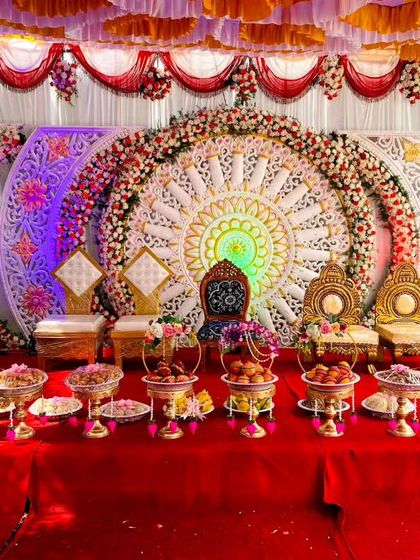 A vibrant and traditional stage setup for a wedding. The backdrop has intricate circular patterns, and the table is lined with my rental platters filled with colorful sweets and offerings.