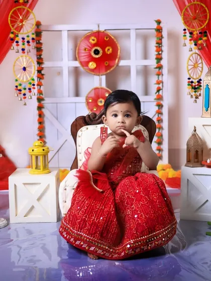 A charming solo portrait of a baby girl during her Diwali photoshoot. Seated on a small chair, she is dressed in a lovely red traditional outfit, surrounded by lanterns and candles in our festive studio setup.