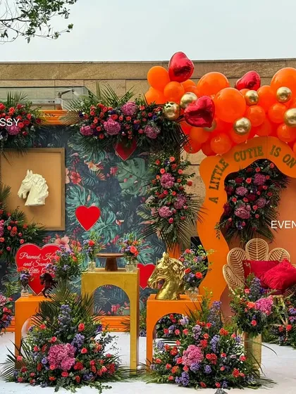 The complete "A Little Cutie Is On The Way" baby shower setup, featuring a lush floral and balloon arch against a tropical print backdrop. The design incorporates a lit-up heart, a peacock chair for the mom-to-be, and a vibrant orange, red, and gold color scheme.