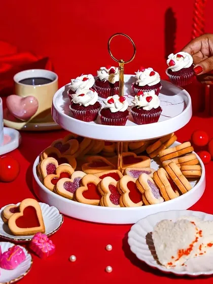 A hand reaches for a red velvet cupcake from a dessert stand laden with Valentine's treats. This action shot adds a dynamic and inviting element to the romantic, red-themed spread.